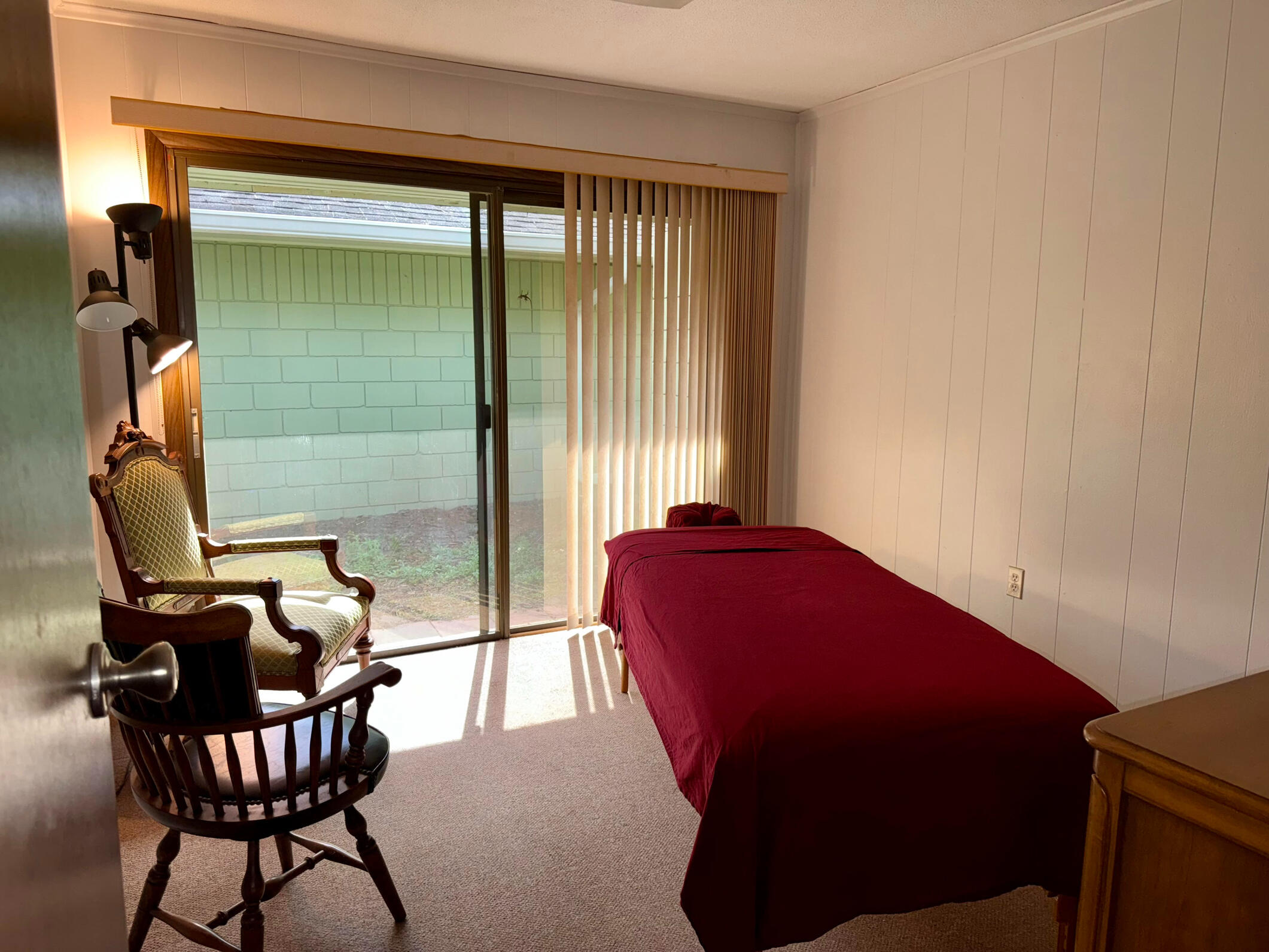 session_space View of the massage room from the door; massage table with red sheets on the right, two chairs on the left. The far wall is mostly taken by a glass door to a private garden.